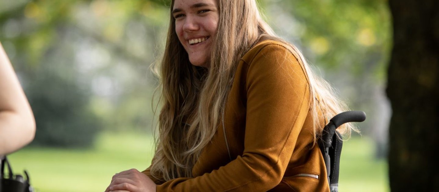 Georgia outside, smiling at the camera. She is in her wheelchair and wearing a tan jacket.