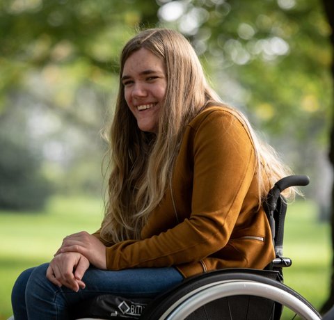 Georgia outside, smiling at the camera. She is in her wheelchair and wearing a tan jacket.