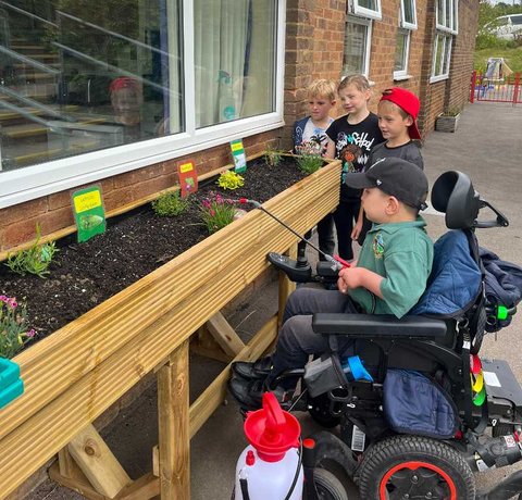 A young wheelchair user waters a flower bed. He is with a group of three other young children who are watching on
