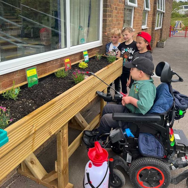 A young wheelchair user waters a flower bed. He is with a group of three other young children who are watching on