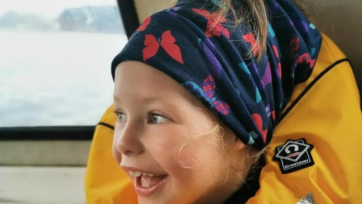 Wren is inside a boat wearing a life jacket. There is a large body of water through the window. She has a huge smile on her face.