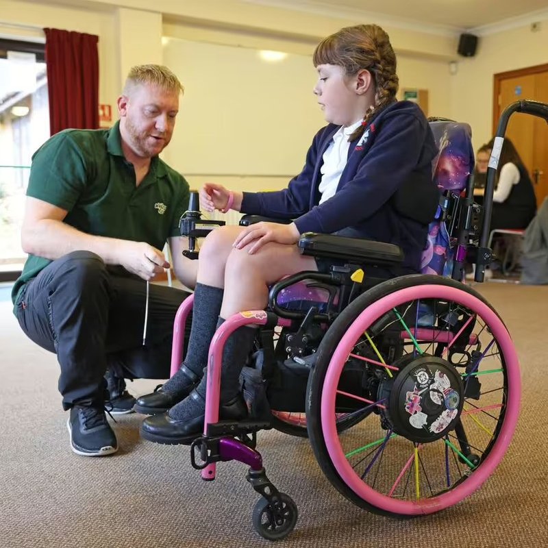Amber is sitting in her pink and purple wheelchair, while a Whizz Kidz engineer is working on it