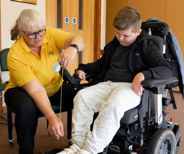 Sarah Wallace in a clinic setting measuring a wheelchair for a young person next to her.