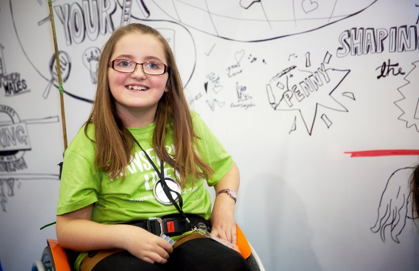 Penny from Kid Board, aged around 7? She is wearing a lime green t-shirt, sitting in her orange wheelchair in front of a white board. She is smiling at the camera. On the whiteboard are doodles and lettering. written