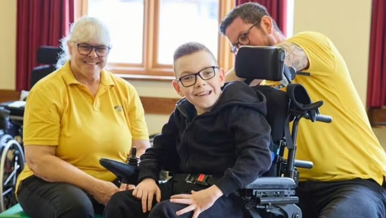Logan has his wheelchair adjusted by two members of Whizz Kidz staff