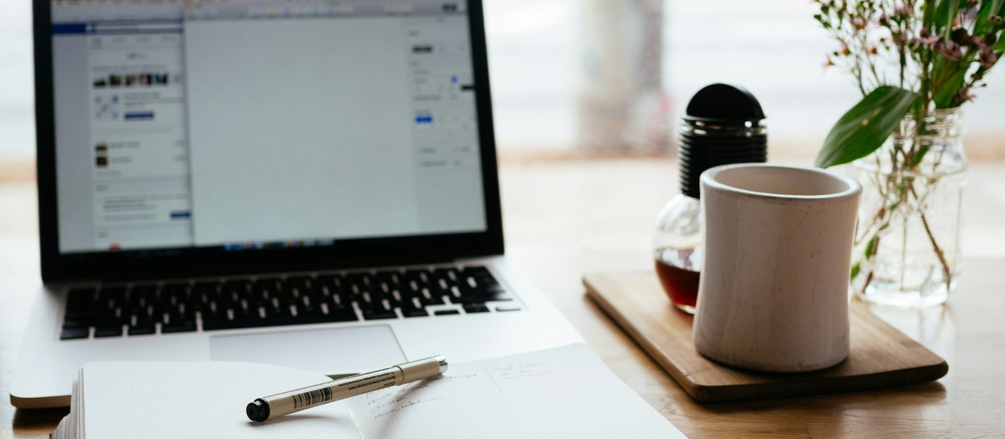 A laptop, an open notepad and a coffee cup sit on a wooden desk
