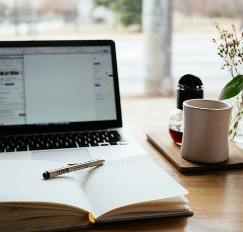 A laptop, an open notepad and a coffee cup sit on a wooden desk