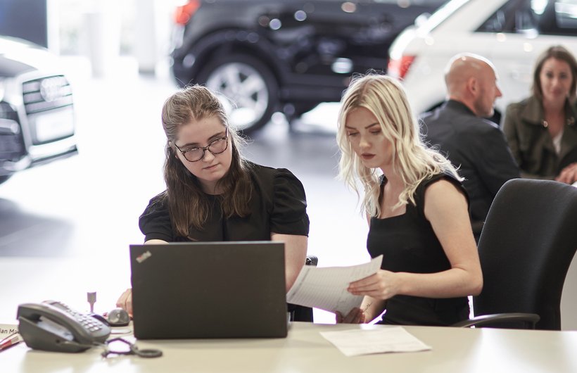 Penelope from Kidz Board sat looking at a laptop. Next to her a woman is showing her a paper document. There are cars in the background.