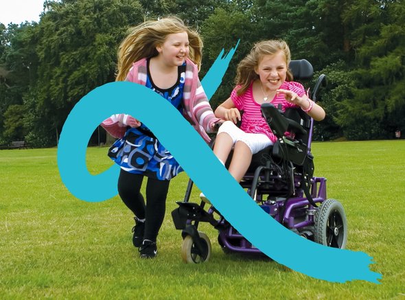 A young wheelchair user plays in her powered chair with her sister running alongside her