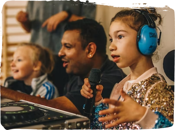 A young child holds a microphone in front of a sound mixing desk. She has an adult to her side