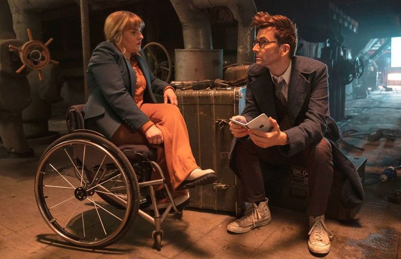 Ruth Madeley as Shirley Anne Bingham in Doctor Who with David Tennant as The Doctor. They are talking in a dark location, with muted lighting. Shirley has crossed her legs.