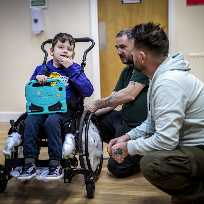 Caius sits in his wheelchair, holding his computer tablet. Next to him are his Dad and a member of Whizz Kidz staff