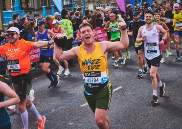 A runner in a yellow Whizz Kidz running vest raises his arms as he goes past a Whizz Kidz cheer point