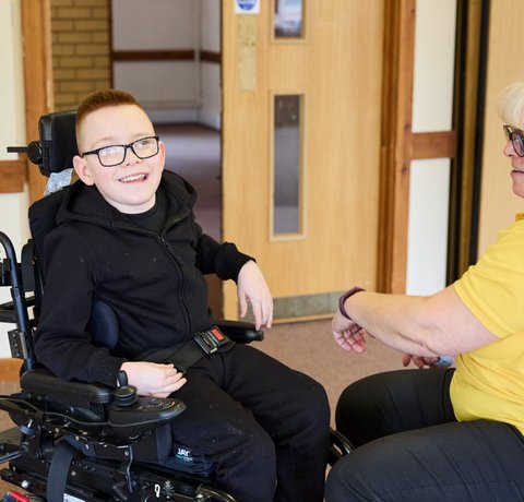 Logan in his wheelchair, smiling for the camera. Sarah Wallace is in front of him, wearing a yellow polo shirt, adjusting his chair.