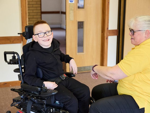 Logan in his wheelchair, smiling for the camera. Sarah Wallace is in front of him, wearing a yellow polo shirt, adjusting his chair.