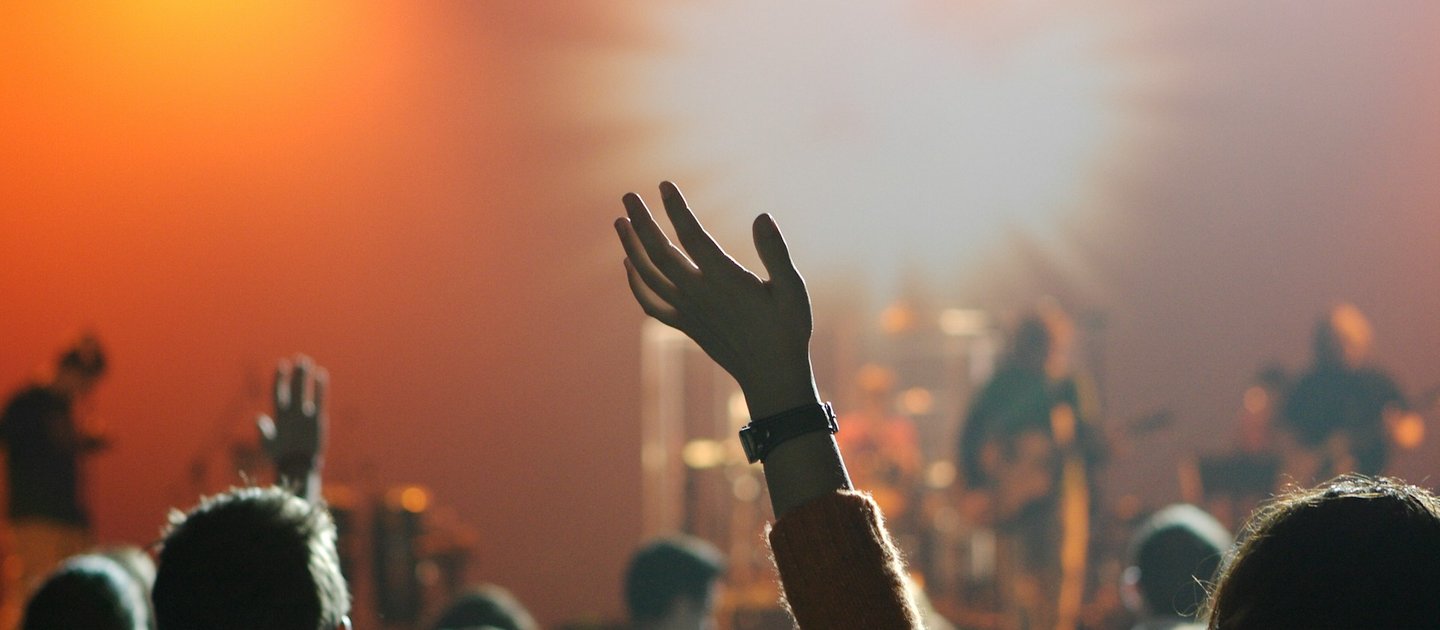A out of focus shot of people enjoying a gig. Someone has their hand up in the air.