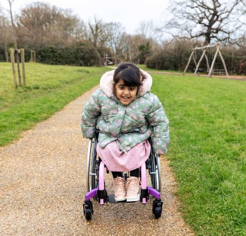 Aniyah outside in her wheelchair whizzing towards the camera. She has a determined look on her face and is wearing a green coat with rainbows on.