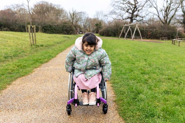 Aniyah outside in her wheelchair whizzing towards the camera. She has a determined look on her face and is wearing a green coat with rainbows on.
