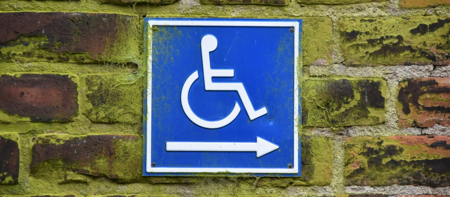 A white wheelchair sign with an arrow pointing right on a blue background. The sign is placed on a brick wall.