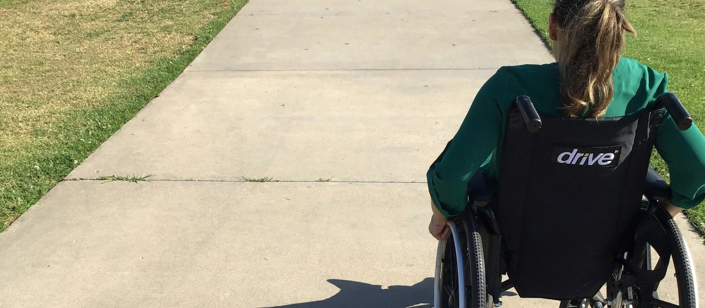 A wheelchair user wearing a green top pushes themselves along a pathway in a park