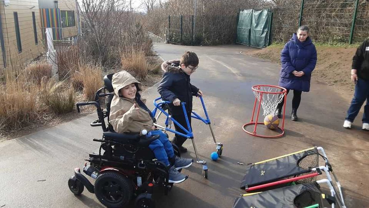 Two young children, one in a powered wheelchair and one with a mobility aid playing with adapted play equipment