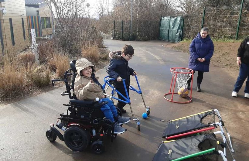 Two young children, one in a powered wheelchair and one with a mobility aid playing with adapted play equipment