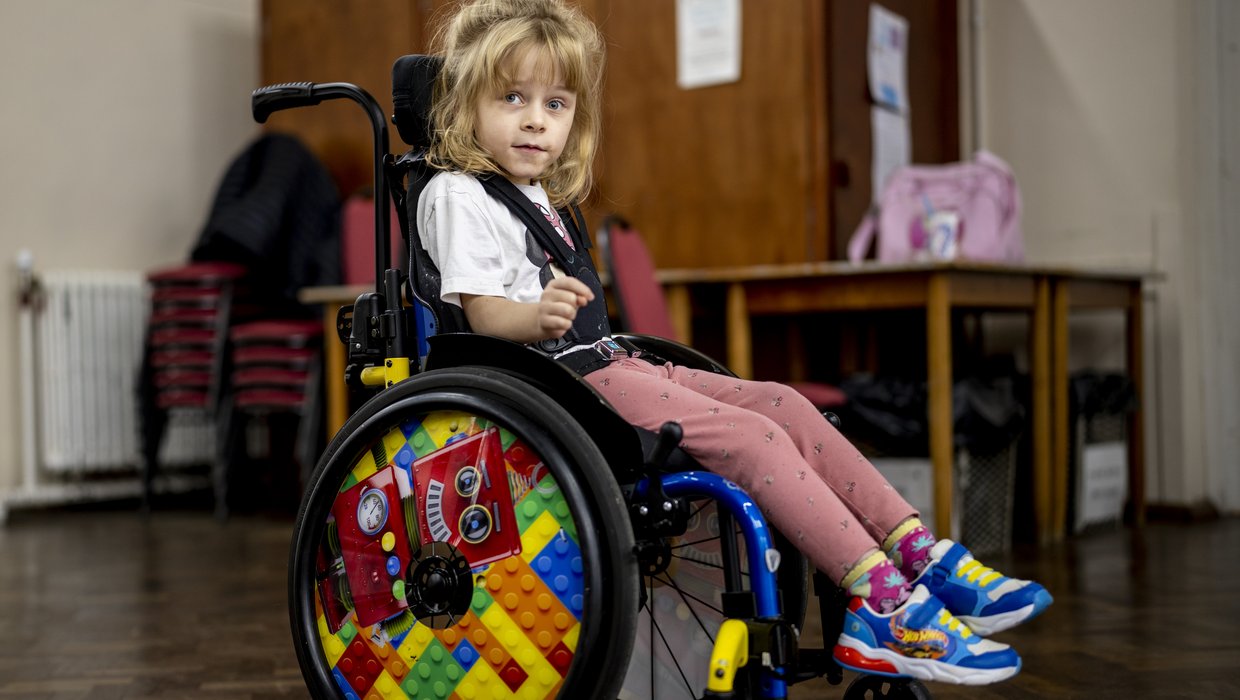 Lucy sits in her wheelchair indoors. Lucy is a small blonde girl, her wheelchair has a lego wheel accessory.