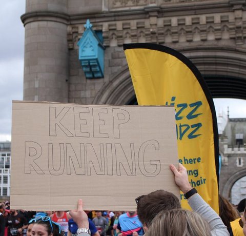 A carboard sign that reads 'keep running' is held up in front of a Whizz Kidz flag