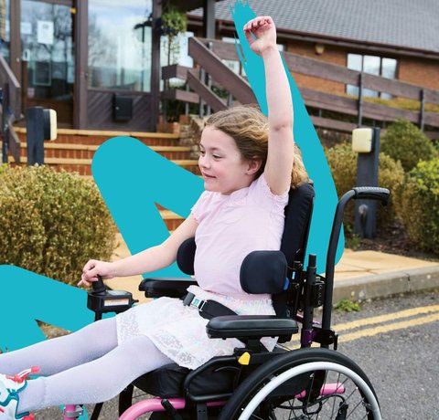 Amber, wearing a pink dress and trainers, whizzes along in her powered chair. Her left arm is raised in the air and the wind is running through her hair.