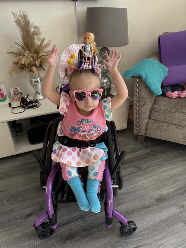 Robyn is sat in her wheelchair, wearing sunglasses indoors. She balances a toy doll, also a wheelchair user, on her head