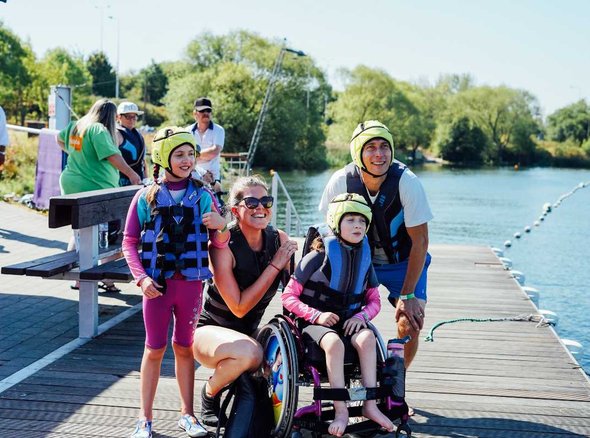 A young wheelchair user with his family grouped together by the side of a large lake
