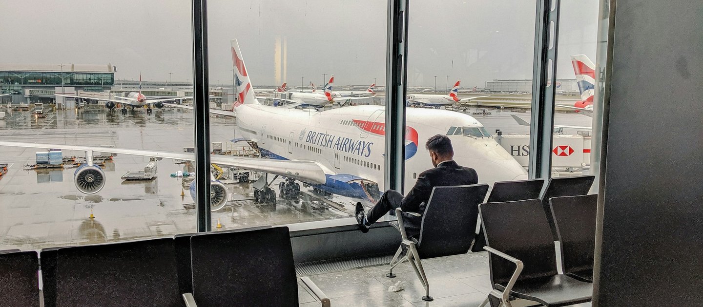 A lone person sits in an airport departure lounge looking out over a British Airways aeroplane in the rain.