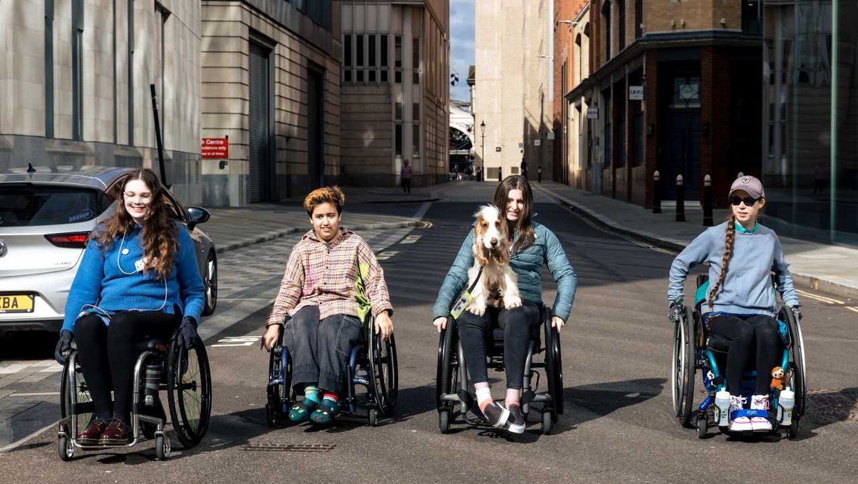 Ruby pushing things forward with other Kidz Board members (from left to right), Caitlin, [Ruby], Ella and Rebecca. They are moving down a street with buildings on both sides. Ella's assistance dog Moose is sat on her lap.