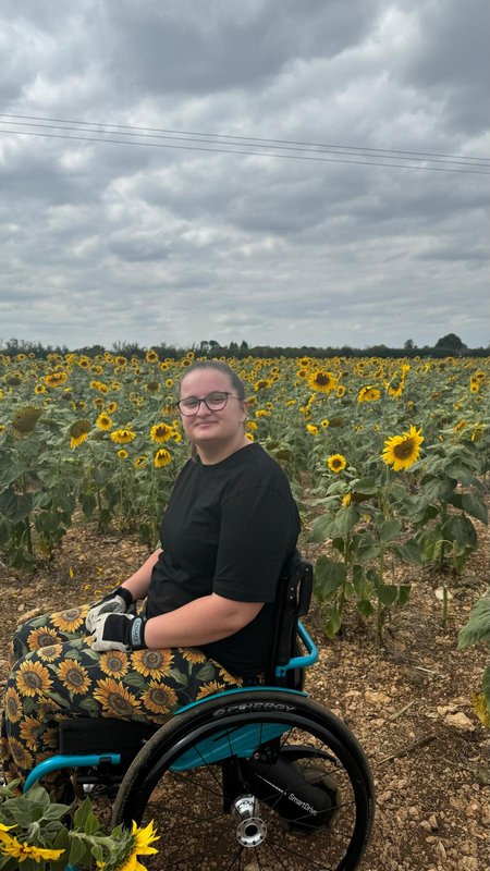 Amber, who is wearing sunflower trousers and glasses, sits in her wheelchair in a field of sunflowers