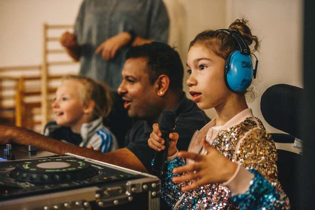 A young wheelchair user sits holding a microphone in front of a sound desk, with other people in the backgroud