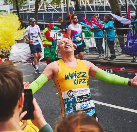 A runner in a yellow Whizz Kidz running vest leans back with his arms out, in awe of the amazing support he is getting at the Whizz Kidz cheer point