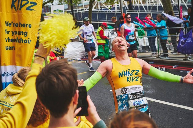 A runner in a yellow Whizz Kidz running vest leans back with his arms out, in awe of the amazing support he is getting at the Whizz Kidz cheer point