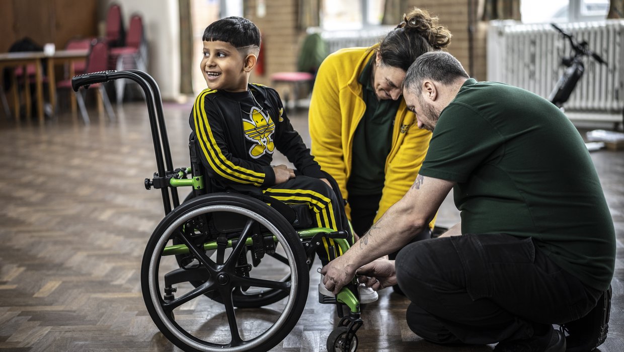 Isaac sits smiling in his wheelchair, while members of the Whizz Kidz team work on the chair.