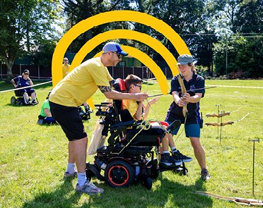 A young wheelchair tries archery. It's outside in a grassy field and the sun is out.