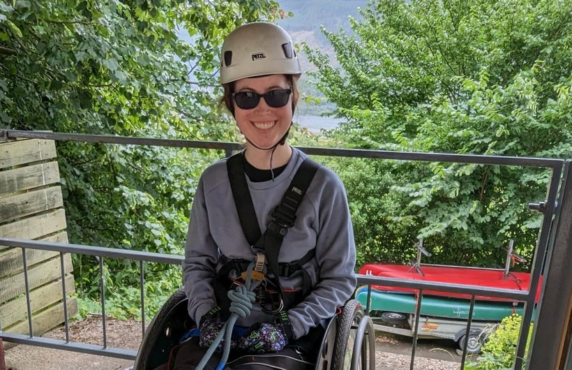 Rebecca from Kidz Board roped up and ready to abseil. She is in her wheelchair, wearing a white climbing helmet and sunglasses.