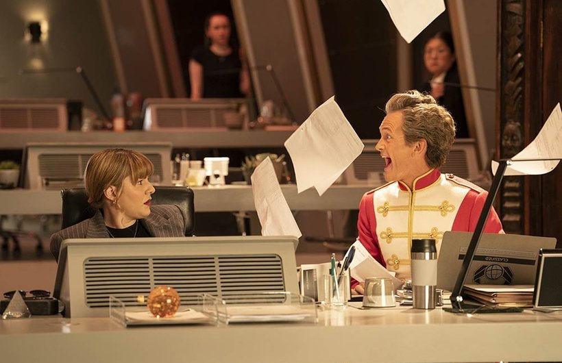 Ruth Madeley as Shirley Bingham behind the desk at UNIT. Neil Patrick Harris as the Toymaker is looking at her shocked face as he scatters papers in the air and laughs.