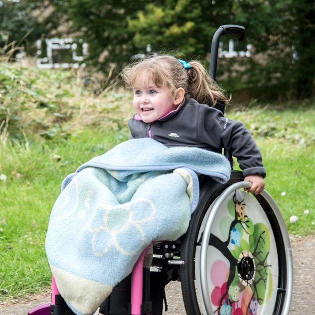 A young wheelchair user pushes herself along a pathway in a park. She has a really cool pink manual wheelchair and a big smile on her face.