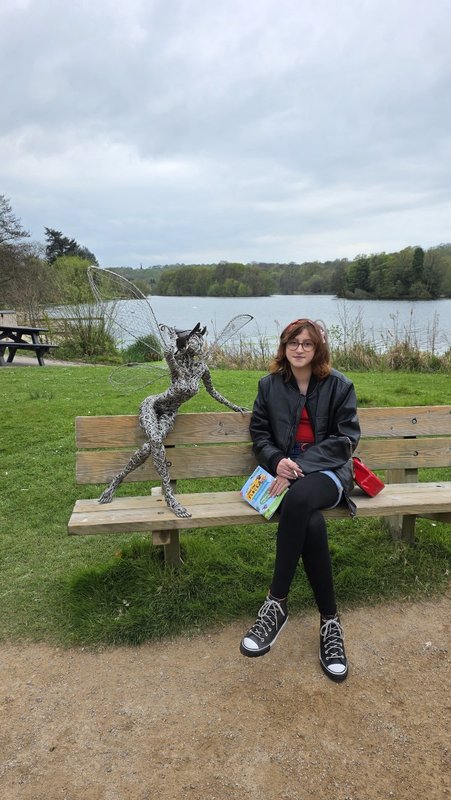Ivy sits on a bench with a book and pencil in her hands. There is a sculpture next to here and a large lake in the background.