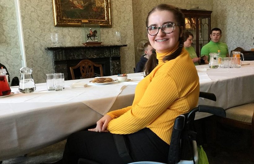 Penelope Harrison half-turning to look at the camera and smile. She is wearing a bright yellow top. She is sat at a long dining table in a formal looking room with a fireplace.