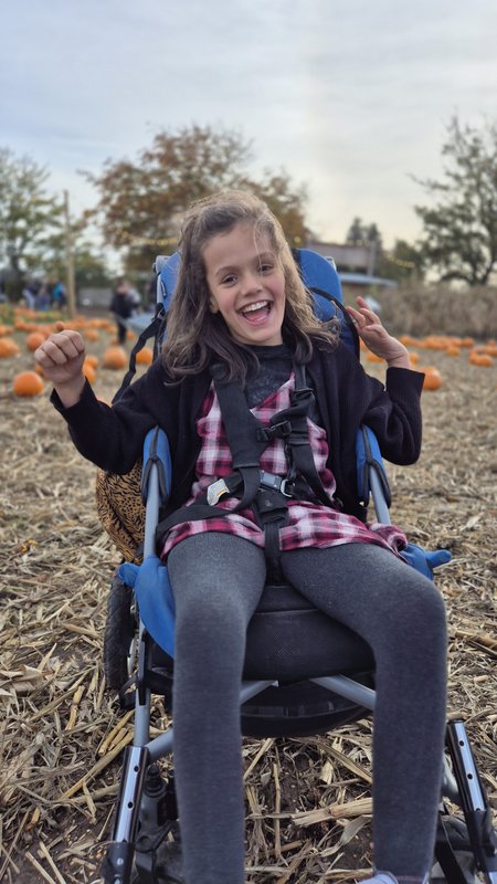 Summer is sitting in a blue buggy, smiling vibrantly at the camera. Behind her you can see a pumpkin patch.