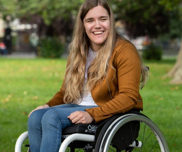 A young wheelchair user sits smiling in a park