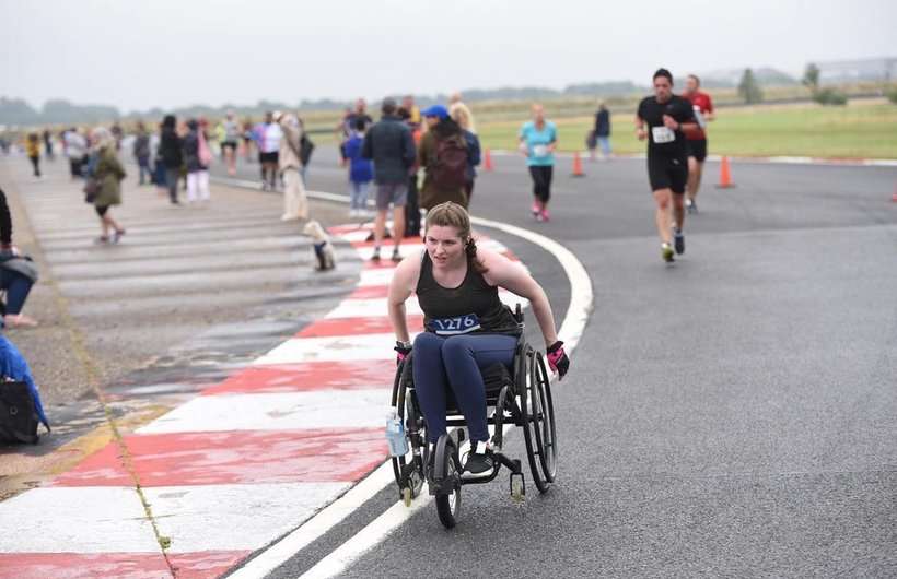 Ella pushing her manual wheelchair around the corner of a race track. There are runners in the background and spectators to the side.