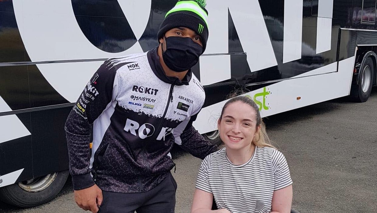 Caitlyn with a race driver posing for the camera. He is wearing his racing suit and a face mask, standing. Caitlyn is in her wheelchair, smiling at the camera