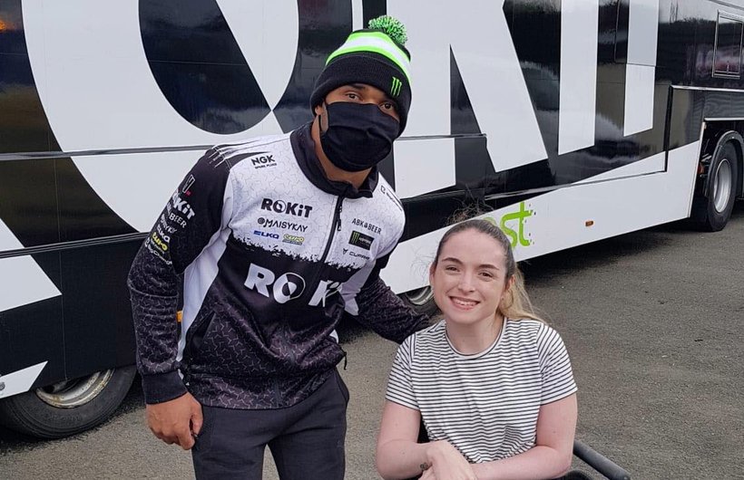 Caitlyn with a race driver posing for the camera. He is wearing his racing suit and a face mask, standing. Caitlyn is in her wheelchair, smiling at the camera