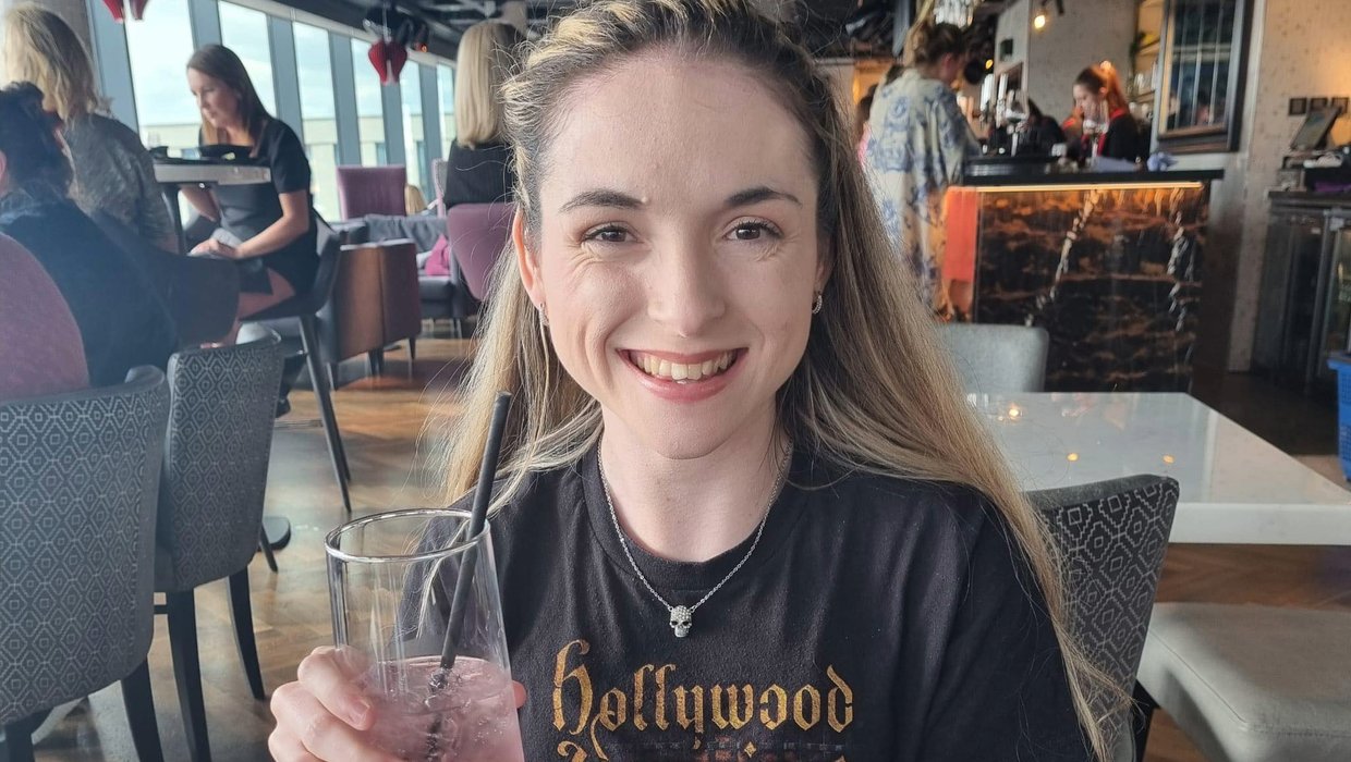 Caitlyn in a bar, smiling at the camera. She is holding a drink and wearing a Hollywood Vampires band t shirt.